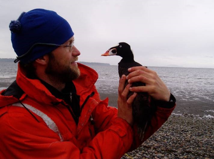 Eric scoter wearing a blue toque and red jacket, holding a shore bird with the ocean in the background and rocky beach