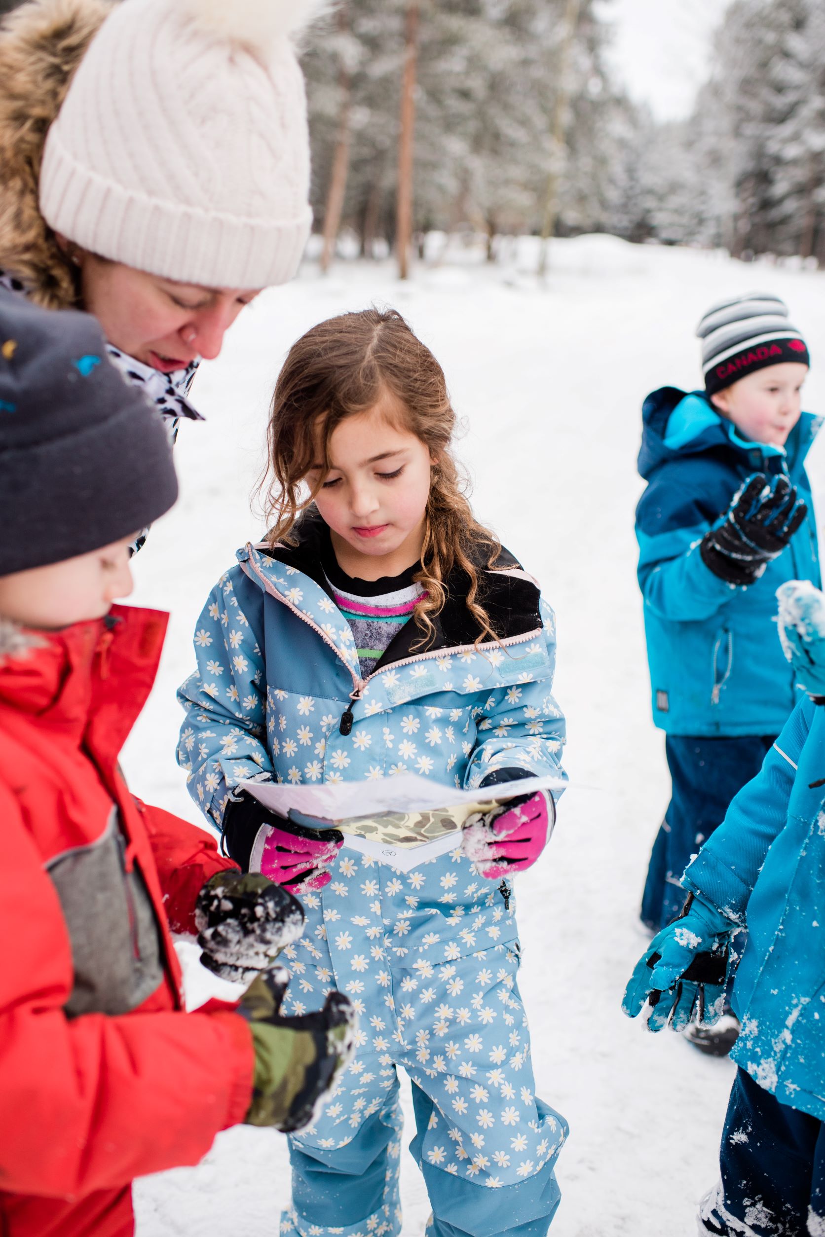 kids looking at a wilderness tracks worksheet to identify animal tracks in the snow, with snowy forest in the background