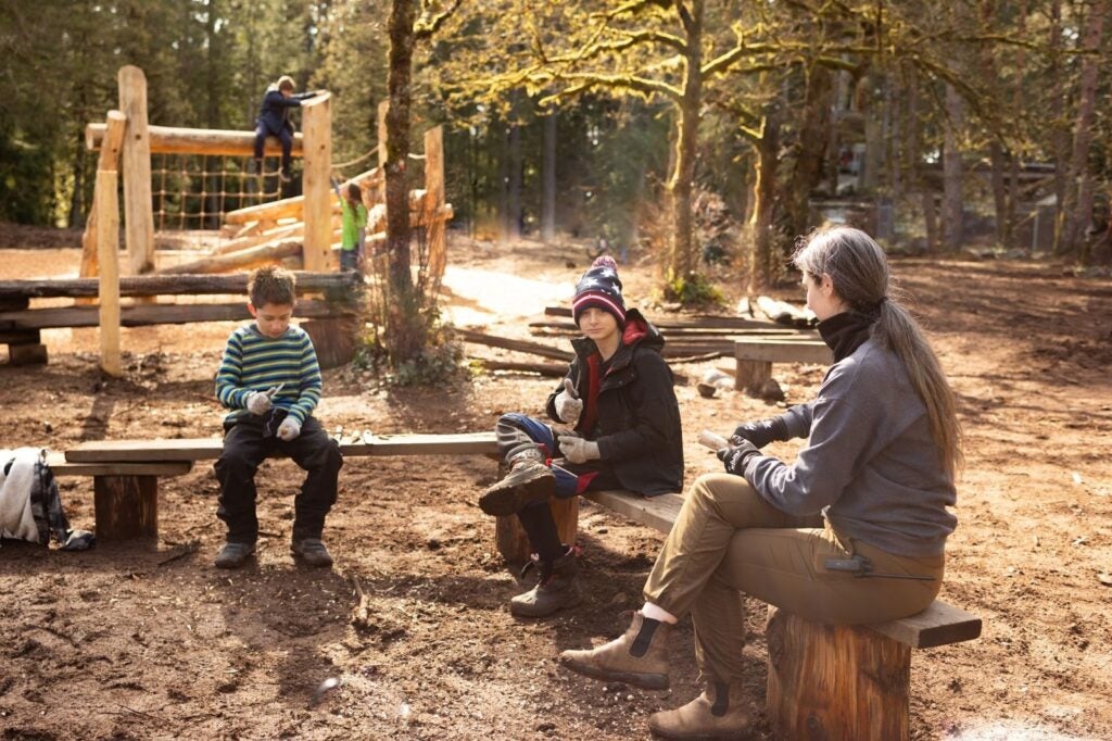 two kids sitting on wooden benches outside learning how to widdle, with the help of an adult leader at wild and immersive