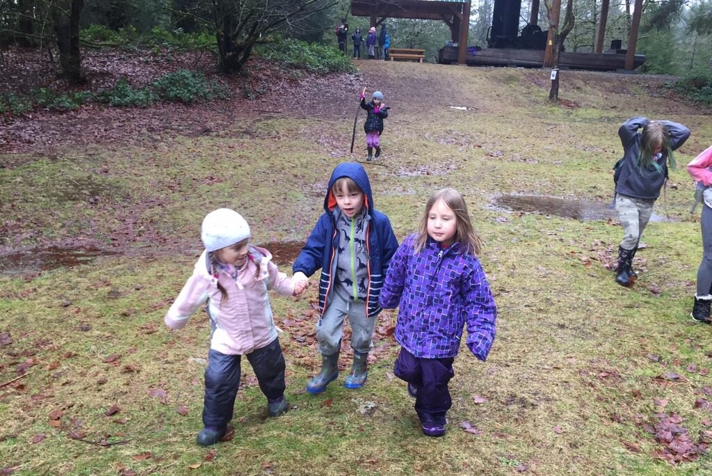 three children holding hands and walking towards the camera on a rainy day, playing outdoors with rain jackets and waterproof pants and rain boots