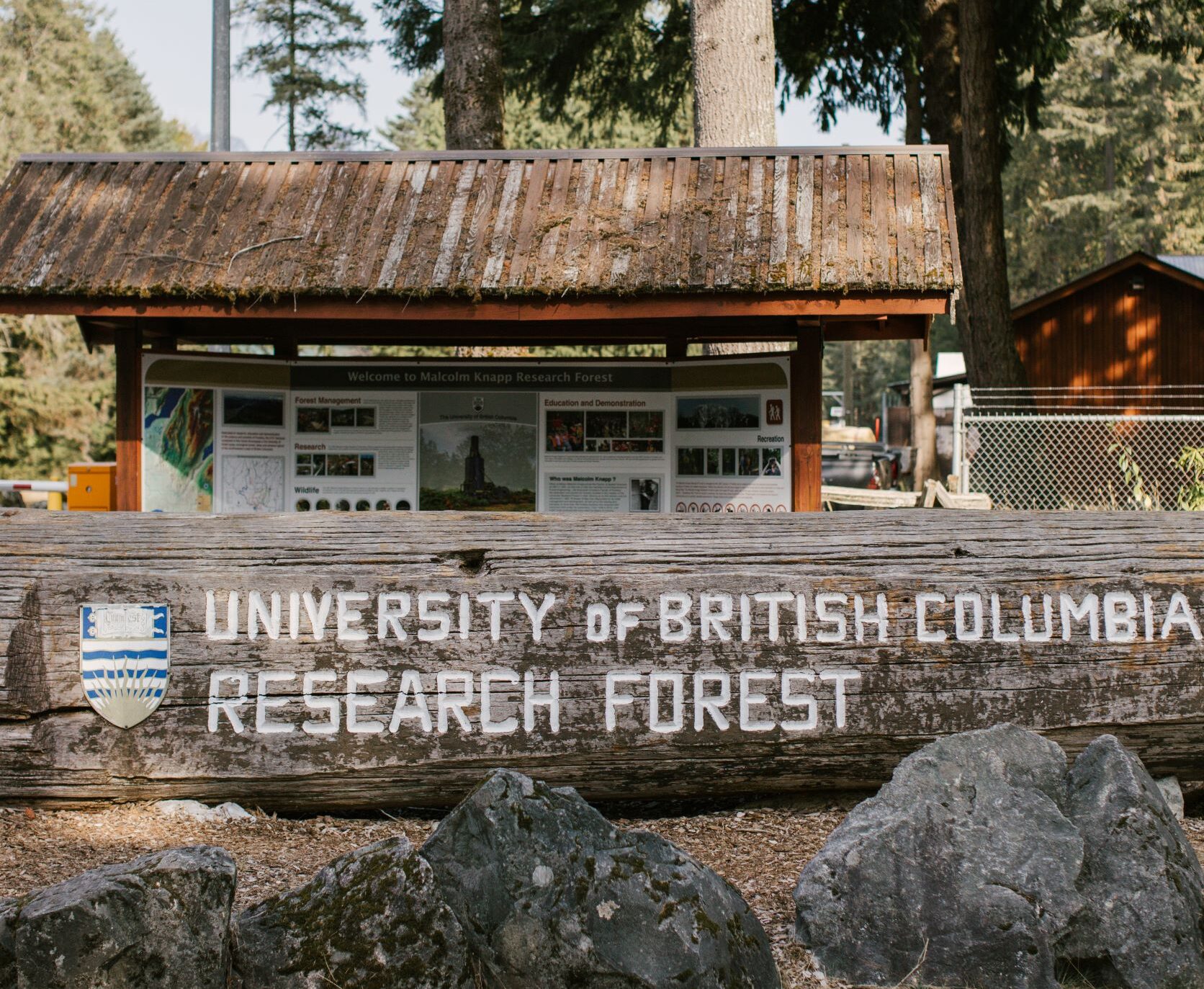 a fallen tree trunk that has been converted in a sign for the University of British Columbia research forest with UBC logo, in front of a sign board that outlines the forest and research