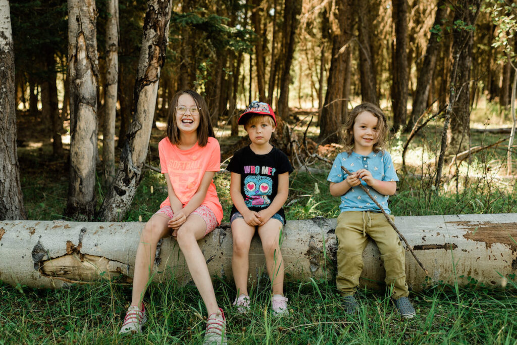 Three young kids sitting on log smiling at camera at a Wild and Immersive Forest Camp on a sunny day, gaining the benefits of outdoor learning