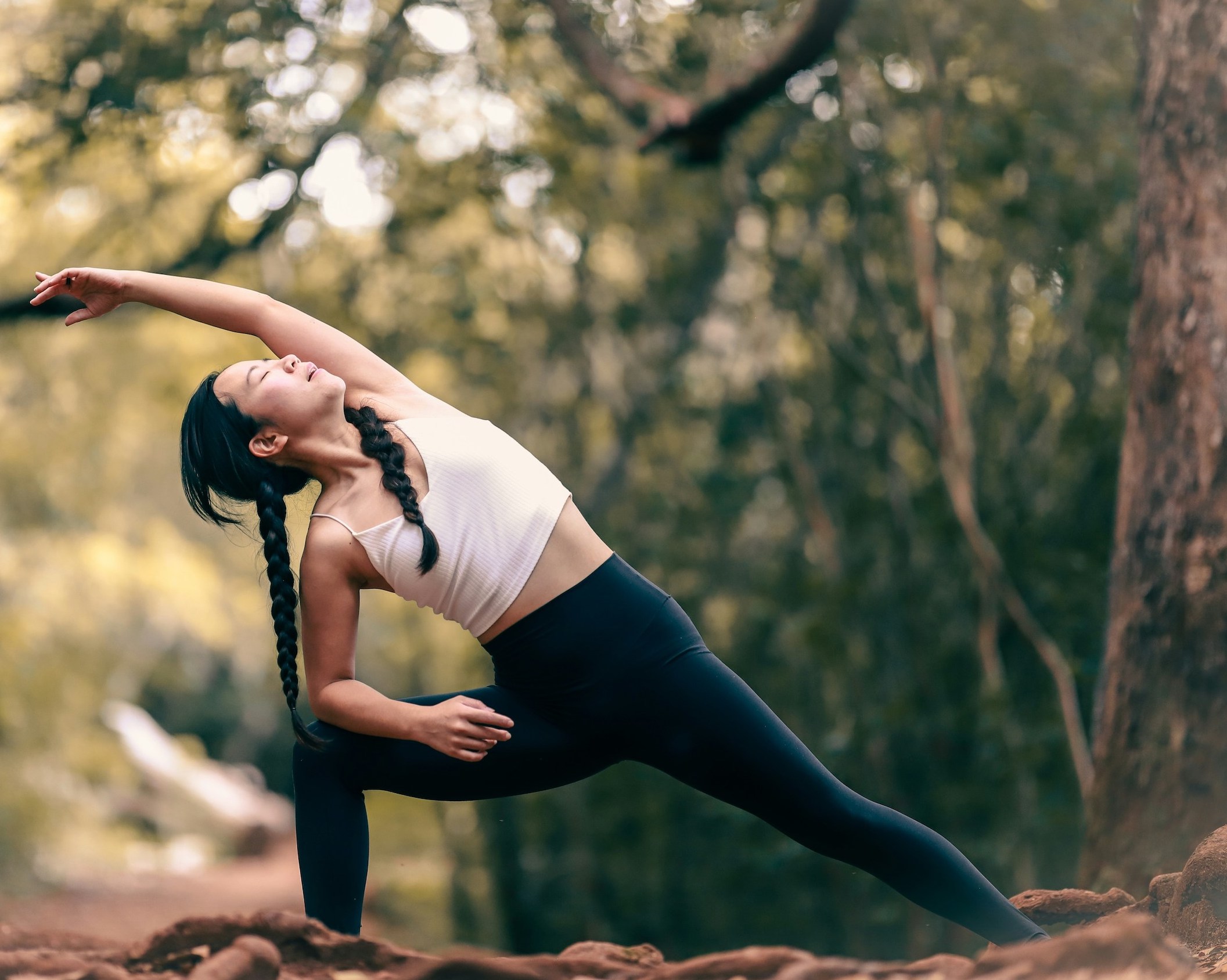 A woman is doing outdoor yoga in dark pants and a light tank top.